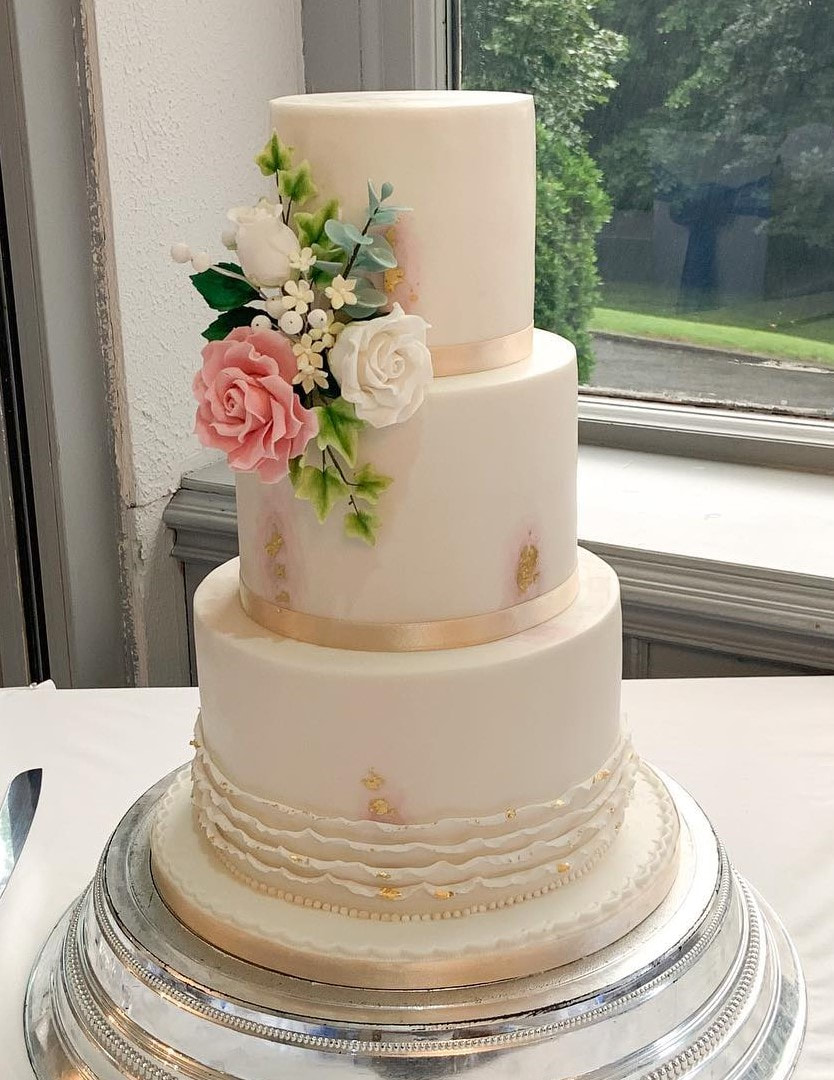 an elegant 3 tier wedding cake with a watercolour effect pattern and decorated with elegant ruffles and of pink and white roses with ivy and green foliage and white berries and blossoms 